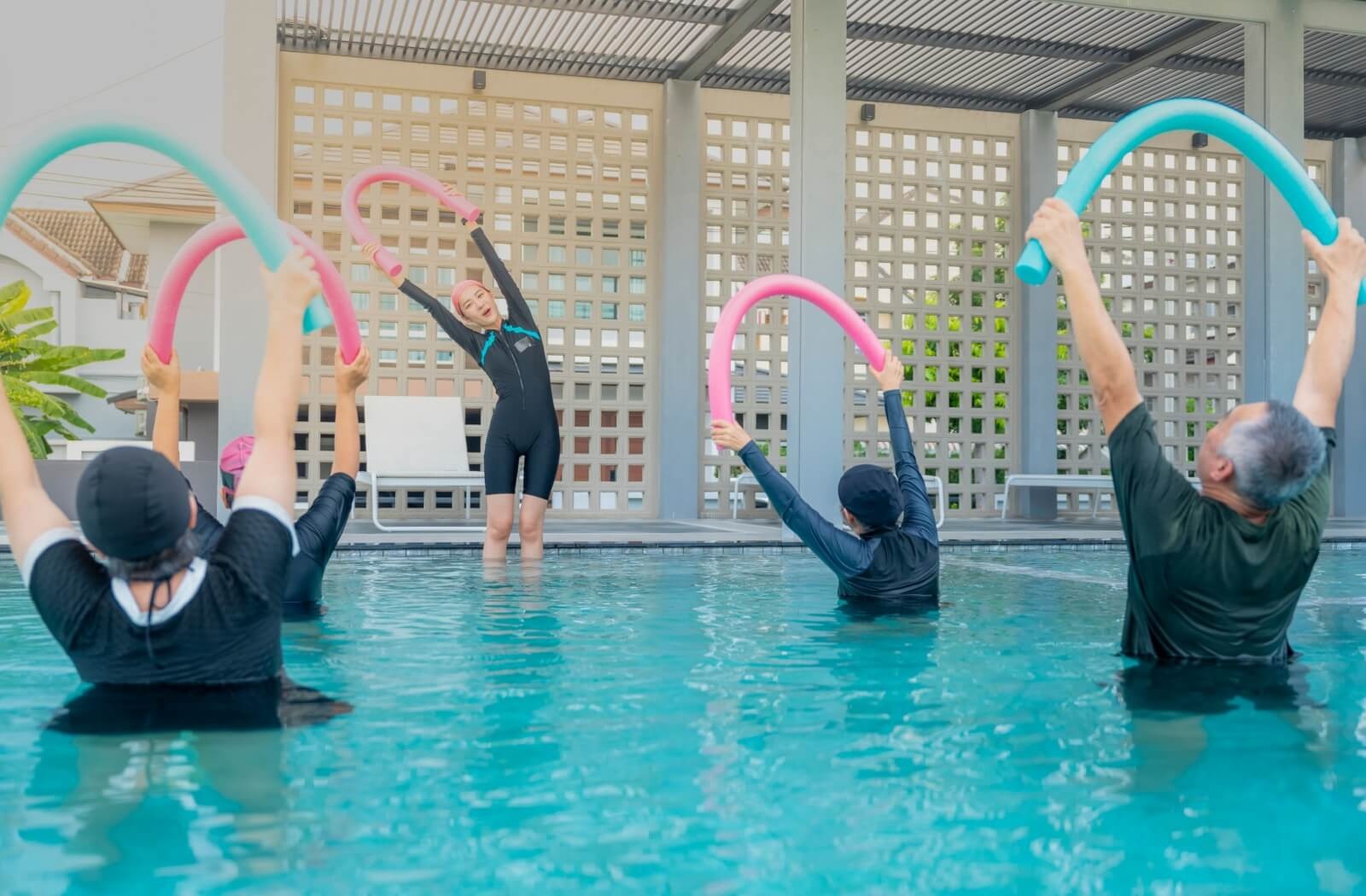 A group of seniors in the shallow end of a pool follow their instructor’s guidance to hold a pool noodle over their heads