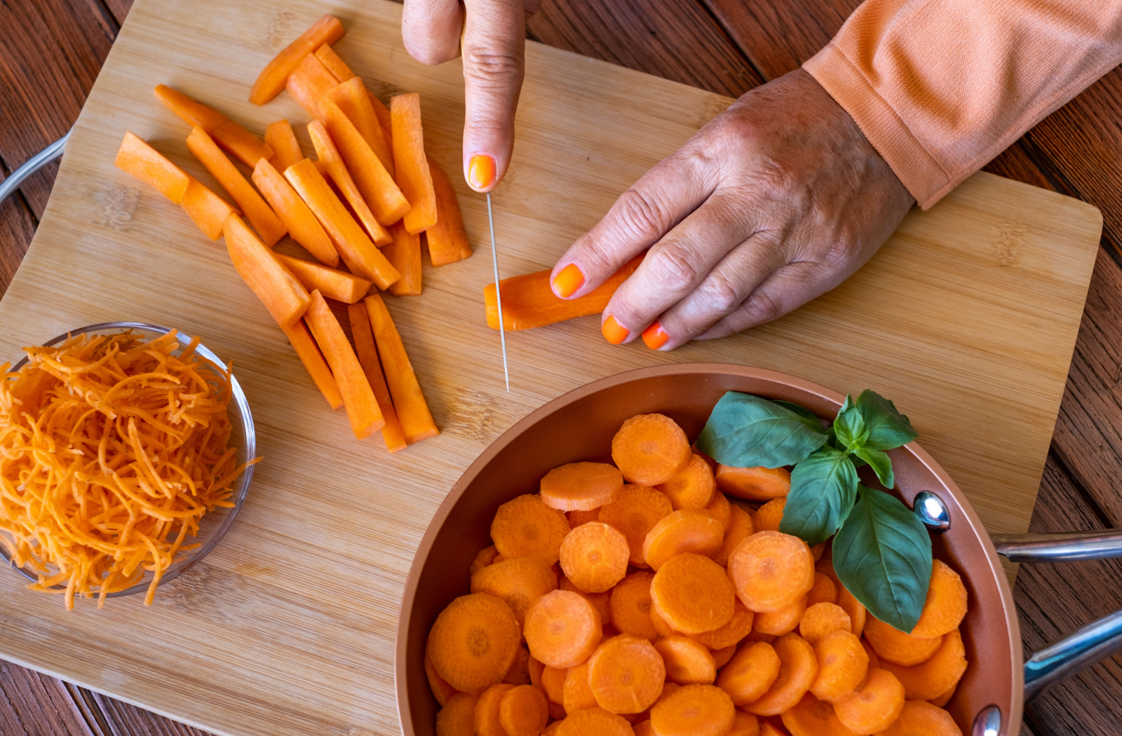A senior slices carrot wedges on a wooden chopping board next to a bowl of shredded carrots and a saucepan with rondelle-cut carrots.