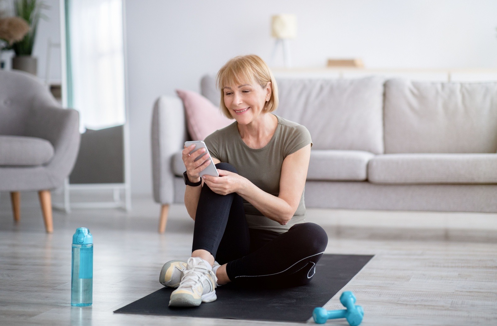 A senior uses their phone while sitting on a yoga mat in their living room.
