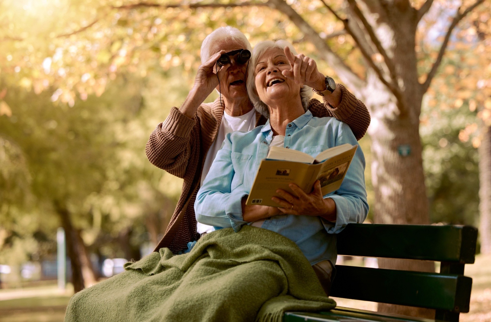 A senior couple spots birds together while sitting on a park bench.
