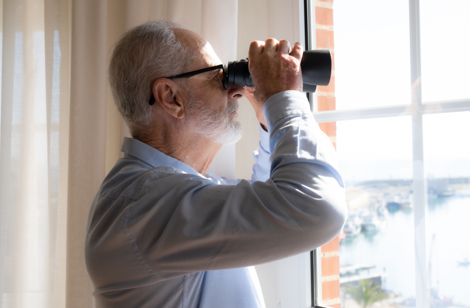 A senior watches a flock of birds through a window using a pair of binoculars.